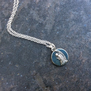 May include: A silver chain necklace with a round pendant featuring a silver angel with wings. The pendant has a dark blue background.