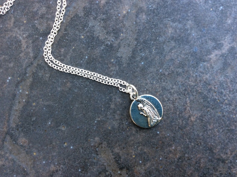 May include: A silver chain necklace with a round pendant featuring a silver angel with wings. The pendant has a dark blue background.