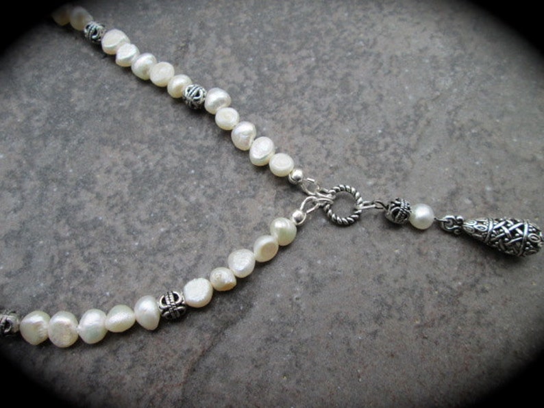 May include: A white pearl necklace with silver accents and a silver pendant. The pendant is shaped like a teardrop and has a decorative design.