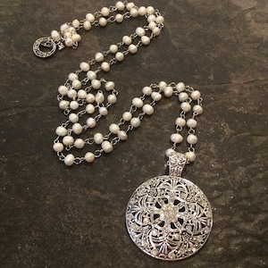 May include: A long silver chain necklace with a large silver pendant featuring a filigree design and clear rhinestones. The necklace is strung with white pearls.