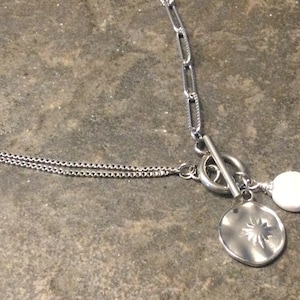 May include: A silver chain necklace with a toggle clasp and two charms. One charm is a round, white bead and the other is a silver disc with a star design.