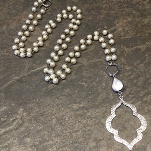 May include: A long white pearl necklace with a silver pendant featuring a teardrop-shaped stone and a decorative openwork design.