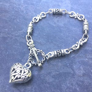 May include: A silver-toned metal bracelet featuring a series of infinity-shaped links and cylindrical beads with engraved patterns. The bracelet is secured with a toggle clasp and includes a large, hollow heart-shaped charm with intricate cutouts.