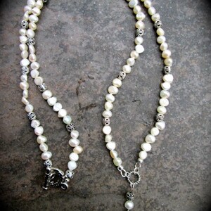 May include: A white pearl necklace with silver accents and a silver pendant. The pendant is shaped like a teardrop and has a detailed design.