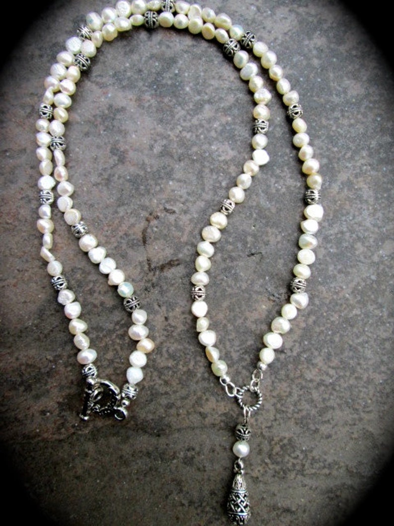 May include: A white pearl necklace with silver accents and a silver pendant. The pendant is shaped like a teardrop and has a detailed design.
