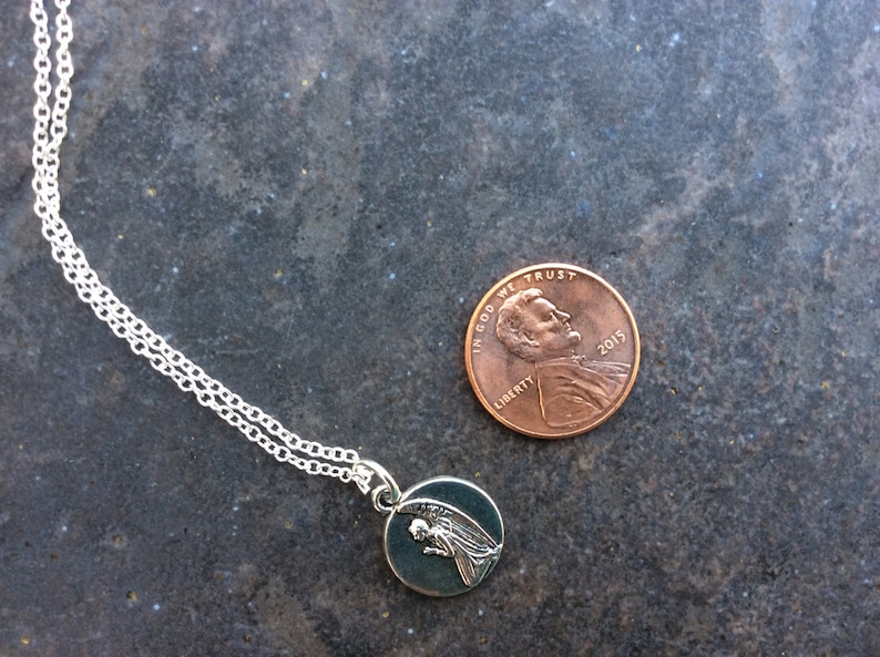 May include: A silver chain necklace with a small round pendant featuring a silver angel with wings. The pendant is about the size of a UK penny.