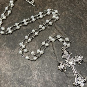 May include: A long silver chain necklace with white faux pearl beads and a large silver cross pendant with rhinestones.