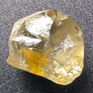 May include: A raw, golden-yellow gemstone with a crystalline structure. The stone has an irregular shape and a polished, reflective surface. The gem appears to be a natural mineral, possibly citrine or topaz, against a dark background.