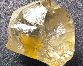 May include: A rough-cut, golden-yellow gemstone with a crystalline structure. The stone has a natural, irregular shape and a slightly translucent appearance, reflecting light. The gem is likely a raw mineral specimen, suitable for jewelry or collection.
