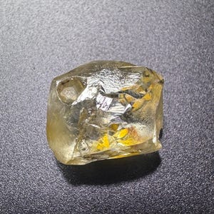May include: A rough-cut, translucent gemstone with a pale yellow hue. The stone has a crystalline structure with multiple facets, reflecting light. The gemstone is set against a dark, textured background, highlighting its natural form and color.