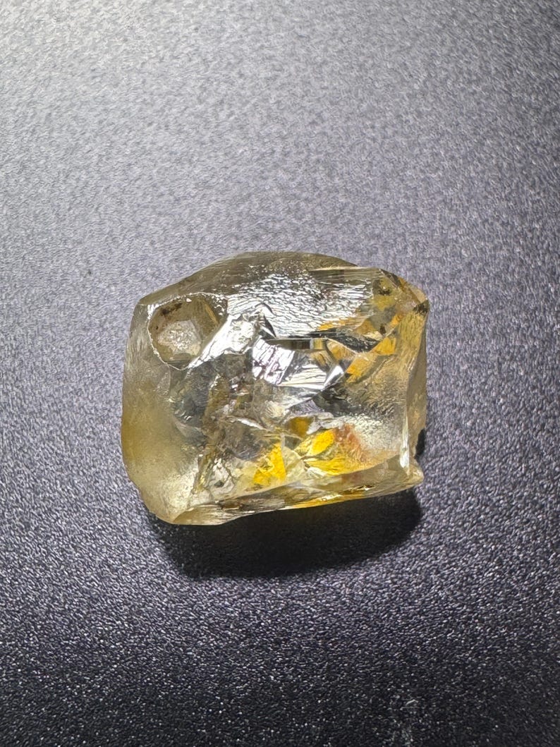 May include: A rough-cut, translucent gemstone with a pale yellow hue. The stone has a crystalline structure with multiple facets, reflecting light. The gemstone is set against a dark, textured background, highlighting its natural form and color.