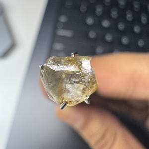 May include: A raw, uncut gemstone ring. The ring features a large, irregularly shaped, translucent yellow-brown stone held in place by silver prongs. The ring is set against a blurred background.