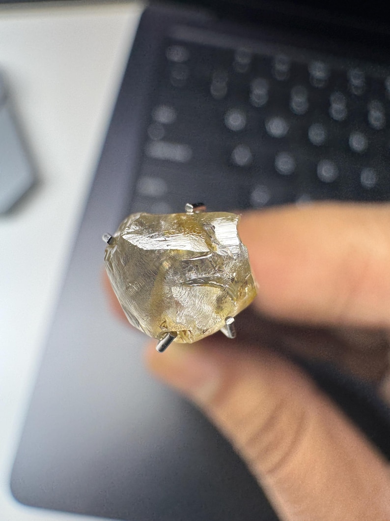 May include: A raw, uncut gemstone ring. The ring features a large, irregularly shaped, translucent yellow-brown stone held in place by silver prongs. The ring is set against a blurred background.