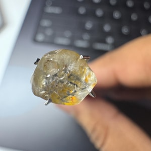 May include: A rough-cut, light yellow gemstone with a silver prong setting. The stone has a cloudy appearance with hints of orange and yellow. The gemstone is held up against a blurred background.