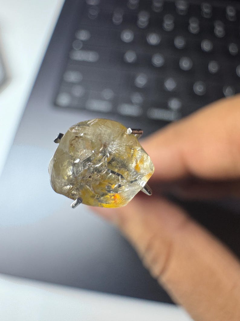 May include: A rough-cut, light yellow gemstone with a silver prong setting. The stone has a cloudy appearance with hints of orange and yellow. The gemstone is held up against a blurred background.
