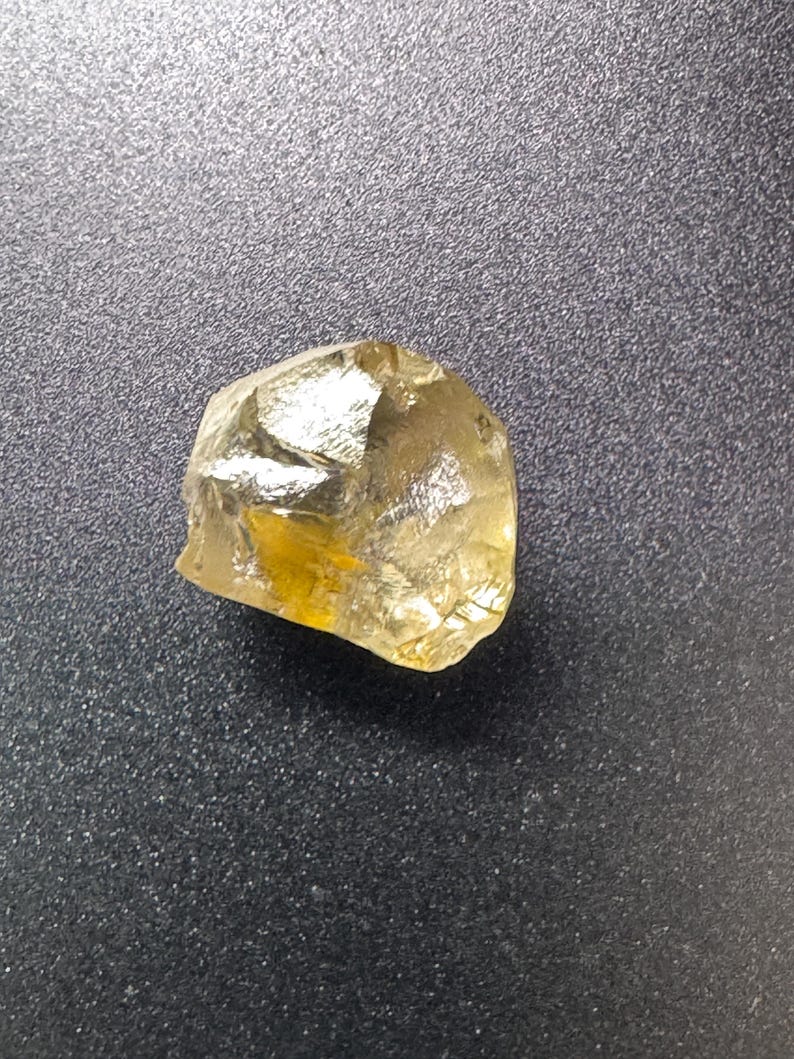 May include: A raw, golden-yellow gemstone with a crystalline structure. The stone has an irregular shape and a polished, reflective surface. The gem appears to be a natural mineral, possibly citrine or topaz, against a dark background.