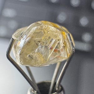 May include: A rough-cut, translucent gemstone with a yellowish hue is held by a silver metal tool. The gem has visible internal structures and facets, reflecting light. The tool's prongs securely cradle the stone, showcasing its natural form.