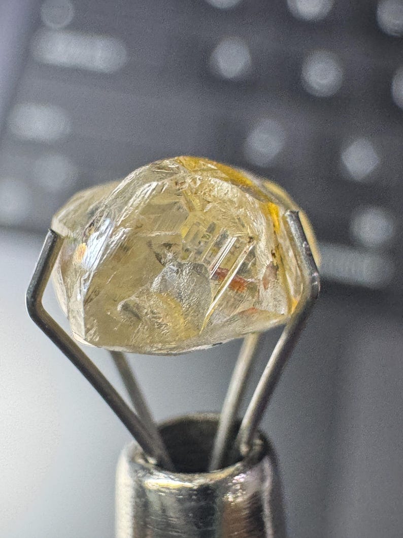 May include: A rough-cut, translucent gemstone with a yellowish hue is held by a silver metal tool. The gem has visible internal structures and facets, reflecting light. The tool's prongs securely cradle the stone, showcasing its natural form.