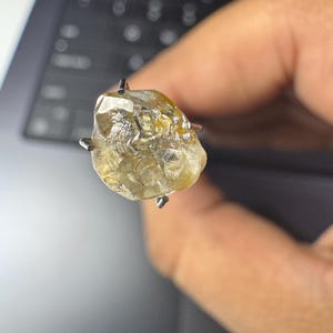May include: A rough-cut, light yellow diamond gemstone held by a four-prong setting. The diamond has a textured surface and is held up against a blurred background.
