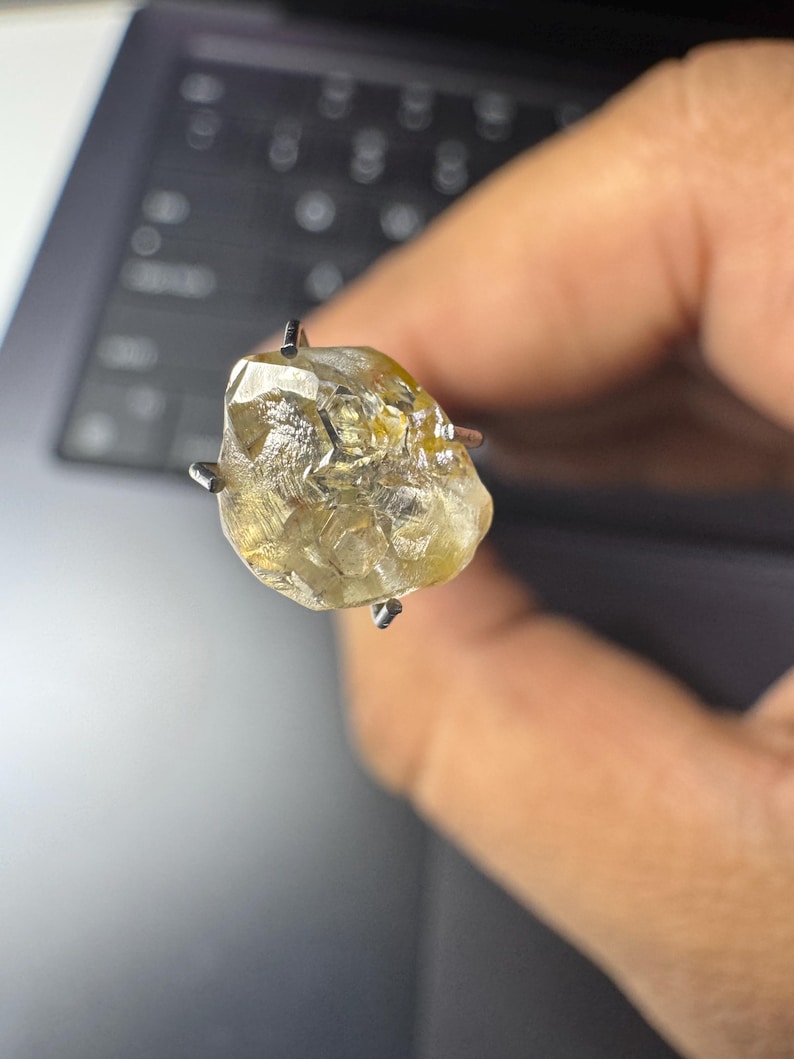 May include: A rough-cut, light yellow diamond gemstone held by a four-prong setting. The diamond has a textured surface and is held up against a blurred background.