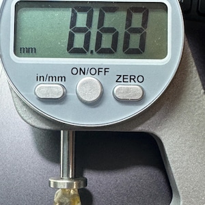 May include: A digital caliper measuring a rough, light-colored gemstone. The display reads 8.68 mm, approximately 0.34 inches. The caliper is silver with black text and buttons. The gemstone is held between the caliper's jaws.