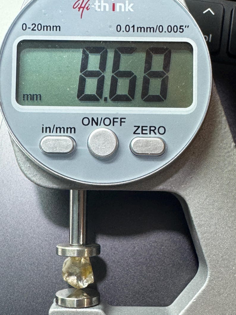 May include: A digital caliper measuring a rough, light-colored gemstone. The display reads 8.68 mm, approximately 0.34 inches. The caliper is silver with black text and buttons. The gemstone is held between the caliper's jaws.