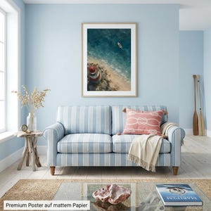May include: A framed vertical wall art print featuring an aerial view of a red and white lighthouse on a sandy beach with a small boat in the blue ocean. The poster is displayed in a light blue room above a striped sofa with a coral throw pillow.