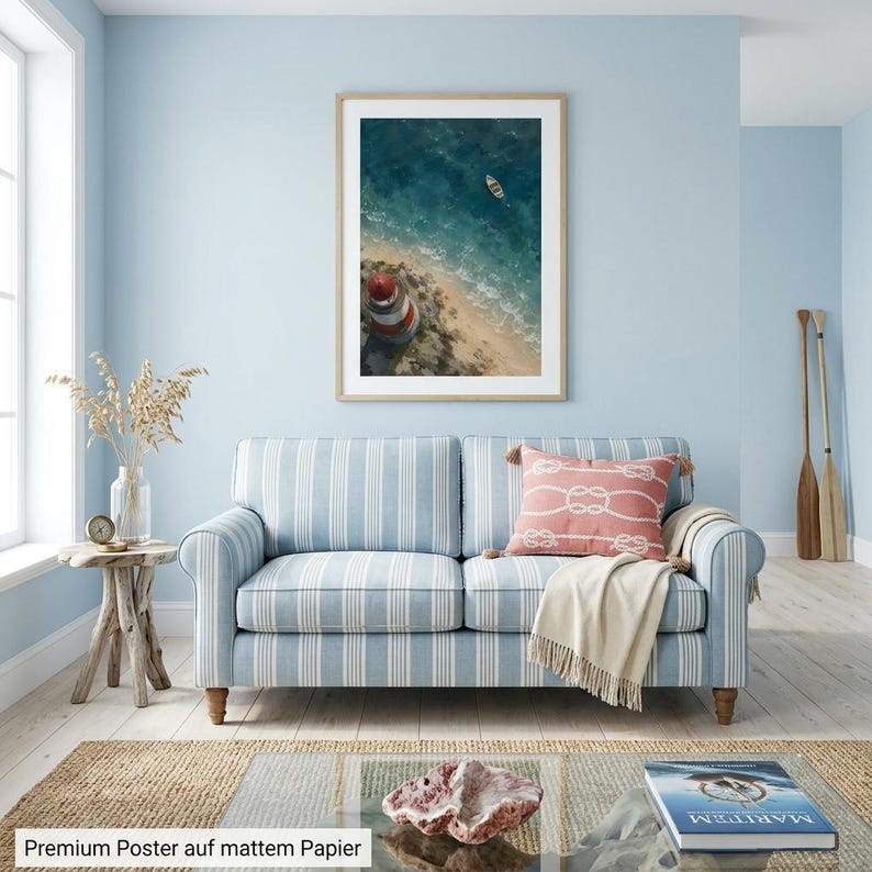 May include: A framed vertical wall art print featuring an aerial view of a red and white lighthouse on a sandy beach with a small boat in the blue ocean. The poster is displayed in a light blue room above a striped sofa with a coral throw pillow.