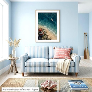 May include: A framed wall art print featuring an aerial view of a red and white lighthouse on a sandy beach with a small boat in the deep blue ocean. The print is displayed above a light blue and white striped sofa with a coral throw pillow.