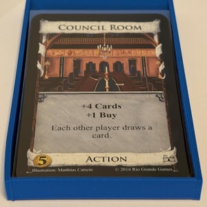 May include: A blue card holder containing a game card labeled "Council Room." The card features an illustration of a room with a chandelier and text indicating card draws and buys. The holder has text that reads "Sleeve-Compatible | Reinforced Walls | Designed to Last."