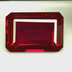 Könnte beinhalten: Ein facettierter, rechteckig geschliffener Rubin-Edelstein. Der Edelstein ist ein tiefes, sattes Rot mit einer hellen, reflektierenden Oberfläche. Der Schliff weist eine Reihe paralleler Facetten auf, die ein brillantes Spiel von Licht und Farbe erzeugen. Auf weißem Hintergrund.