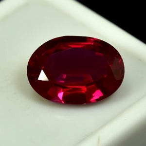Puede incluir: Una gema de rubí facetada de forma ovalada. La gema es de un color rojo intenso y saturado con una superficie brillante y reflectante. La gema está sobre un fondo blanco.