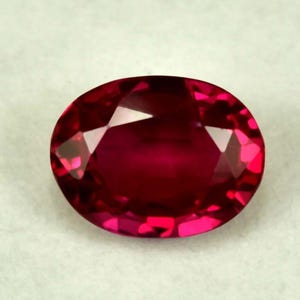 Può includere: Una gemma di rubino sfaccettata a taglio ovale. La gemma è di un colore rosso intenso e saturo, con brillanti riflessi interni. Le sfaccettature creano un gioco di luce sulla superficie, evidenziando la sua chiarezza e brillantezza. La gemma è posta su uno sfondo neutro.