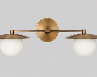 Puede incluir: Un aplique de pared con una barra horizontal de latón unida a una placa trasera circular de latón. Dos globos de cristal esféricos blancos cuelgan bajo pantallas anchas, planas y circulares de latón a ambos lados del soporte central.
