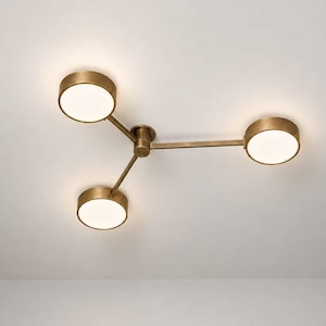 May include: A ceiling light fixture featuring three circular, brass-toned metal housings arranged in a radial pattern. Each housing contains a flat, circular white diffuser that emits a soft, warm glow, connected to a central hub by thin arms.