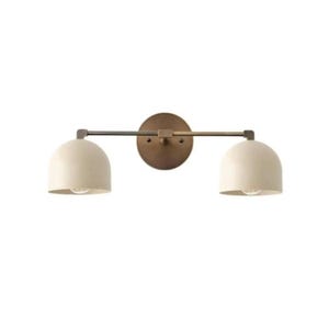 May include: A two-light vanity light fixture with a round, bronze-colored backplate and a horizontal bar. Two off-white, dome-shaped shades are attached to the bar. The fixture has a modern design and is suitable for a bathroom or powder room.