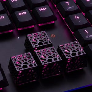 Può includere: Una tastiera per computer nera con tasti standard e tre tasti freccia personalizzati caratterizzati da un intricato design a reticolo traforato. I tasti sono illuminati dal basso con una vivace luce rosa.