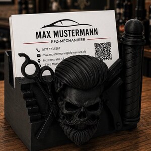 Op de afbeelding: Een zwarte visitekaarthouder met een gebeeldhouwde schedel met een pompadour-kapsel en baard, geflankeerd door een schaar en een kapperspaal. De houder bevat een wit visitekaartje voor Max Mustermann, KFZ-Mechaniker.
