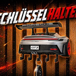 Könnte beinhalten: Silberner Porsche GT3 Schlüsselhalter mit roten Akzenten und einem schwarzen Spoiler. Der Schlüsselhalter hat vier Haken und den Text "SCHLÜSSEL HALTER" und "PORSCHE" im Hintergrund.