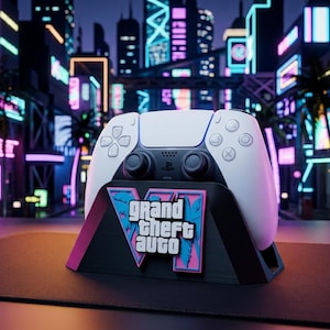 Könnte beinhalten: Ein weiß-schwarzer kabelloser Gamecontroller auf einem schwarzen Ständer. Der Ständer zeigt eine pink-blaue Grafik mit dem Text Grand Theft Auto in weißen Buchstaben. Der Controller verfügt über ein zentrales Touchpad, zwei Analogsticks und Tasten.