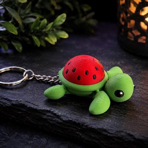 May include: A turtle keyring featuring a bright green body and a red shell patterned with black seeds to resemble a watermelon. The turtle is attached to a silver metal split ring by a short chain, showcasing a smooth, layered 3D printed texture.