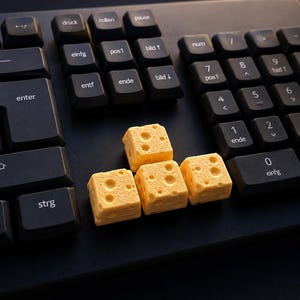 May include: A black computer keyboard featuring three yellow, cube-shaped keycaps designed to resemble Swiss cheese with visible holes. The keyboard includes standard keys with German labels such as enter, strg, entf, and ende, arranged in a standard layout.