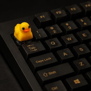 Op de afbeelding: Een zwart computertoetsenbord met een aangepaste gele badeend-toetskap op de tab-toets. Het toetsenbord heeft zwarte toetsen met goudkleurige letters, waaronder de tab-toets, caps lock, shift en diverse lettertoetsen voor tekstinvoer.