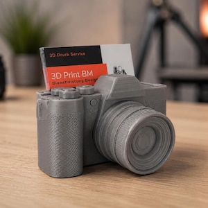Könnte beinhalten: Ein grauer, 3D-gedruckter Visitenkartenhalter in Kameraform mit texturierter Oberfläche. Eine weiße Visitenkarte mit schwarzen und orangen Akzenten steckt im oberen Schlitz und zeigt den Text 3D Druck Service sowie 3D Print BM Dienstleistung Design.