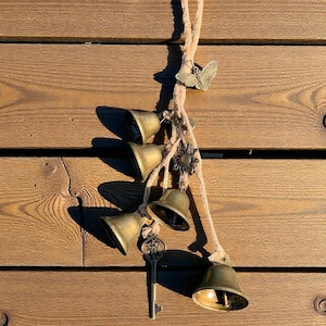 Könnte beinhalten: Dekorative Aufhängung mit fünf Messingglocken, einem Schmetterling, einer Blume und einem Schlüssel. Die Glocken sind an einem Naturseil vor einem Holzgrund befestigt. Die Glocken sind in verschiedenen Größen und Formen.