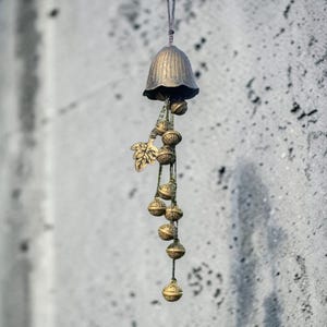 Könnte beinhalten: Eine bronzefarbene Windglocke mit glockenförmigem Oberteil und hängenden kugelförmigen Ornamenten. Ein dekoratives Blatt ist an der Glocke befestigt. Die Windglocke ist an einer dünnen Schnur vor einem strukturierten grauen Hintergrund aufgehängt.