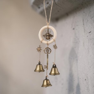Könnte beinhalten: Ein dekorativer Windspiel mit einem Holzring, einer Bronzebiene, einer Sonne, einem Schlüssel und Glocken. Das Windspiel wird von einer natürlichen Schnur aufgehängt. Die Glocken und Charms sind bronzefarben.