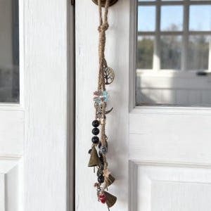 May include: A decorative hanging ornament made from natural jute rope, adorned with charms. These include a tree of life pendant, a floral bead, a crescent moon, black beads, small brass bells, and a red heart. The ornament hangs against a white door.