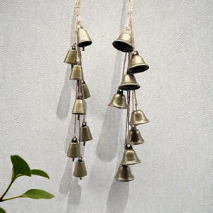 May include: Two strands of bronze-colored bells hanging against a light gray wall. Each strand has multiple bells of varying sizes, strung on natural-colored twine. The bells have a rustic, aged appearance, suggesting a vintage or handcrafted style. The bells are likely used for decoration.