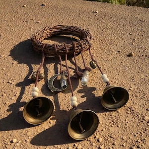 May include: A rustic wind chime featuring a woven grapevine wreath. Brown leather cords suspend brass bells, small glass vials, wooden beads, and a bronze crescent moon charm. The chime is designed for outdoor or indoor decor.